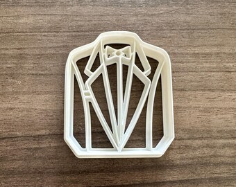 Puede incluir: Cortador de galletas de plástico blanco con forma de chaqueta de esmoquin con pajarita. El cortador tiene forma rectangular con líneas detalladas que representan las solapas, el cuello y la pajarita de la chaqueta. Ideal para galletas festivas.