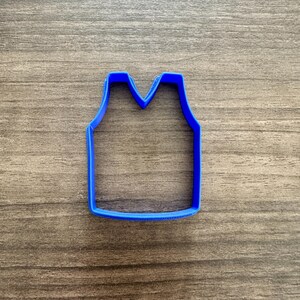 May include: A blue plastic cookie cutter in the shape of a tank top. The cutter has a simple design with a V-neck and a rectangular body. The cutter is made of a solid blue plastic and is sitting on a wooden surface.