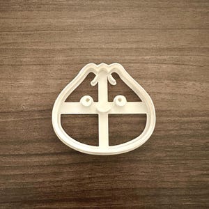 May include: A white plastic cookie cutter in the shape of a dumpling. The cutter has a rounded top with a small handle and two eye-like circles. A cross-shaped design is in the center.