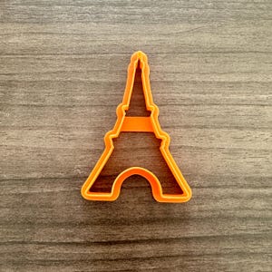 May include: Orange plastic Eiffel Tower cookie cutter. The cutter is a silhouette of the famous Parisian landmark, with a detailed outline of the tower's structure. The cutter is designed for making cookies or other treats in the shape of the Eiffel Tower.