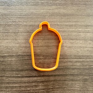 May include: An orange plastic cupcake-shaped cookie cutter. The cutter is a single piece with a rounded top and a flared base. The cutter is sitting on a wooden surface.