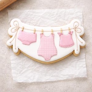 May include: A decorated sugar cookie in the shape of a clothesline with baby clothes. The cookie features a pink diaper, a pink gingham onesie, and a pink top, all hanging from a string with tiny clothespins. The cookie is outlined in white icing.