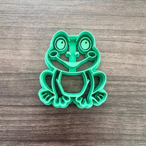 May include: A bright green frog-shaped cookie cutter. The cutter features a detailed outline of a frog with large eyes, a smiling mouth, and webbed feet. The design is suitable for making cookies or other treats.