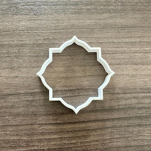 May include: A white, geometric cookie cutter with a decorative, eight-pointed star shape. The cutter is made of plastic and is set against a wooden background. This item is ideal for baking and crafting.