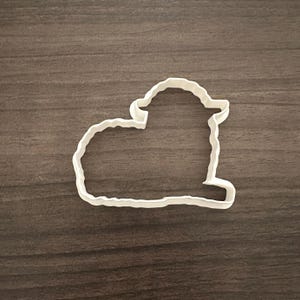 May include: A white plastic cookie cutter in the shape of a lamb, lying down. The cutter has a simple outline with a scalloped edge, and is set against a brown wooden surface.