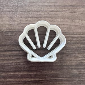May include: A white plastic seashell-shaped cookie cutter. The cutter has a detailed design with curved lines and a scalloped edge, perfect for creating ocean-themed treats. The cutter is set against a dark wood grain background.