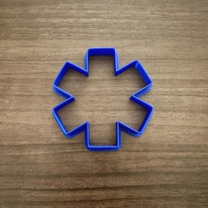 May include: A blue plastic cookie cutter in the shape of a six-pointed star. The cutter is made of a solid blue plastic and is designed to cut through dough or other soft materials. The cutter is on a wooden surface.