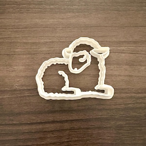 May include: A white plastic cookie cutter in the shape of a lamb, lying down. The cutter has a detailed outline, showing the lamb's head, body, and legs. The cutter is on a brown wooden surface.