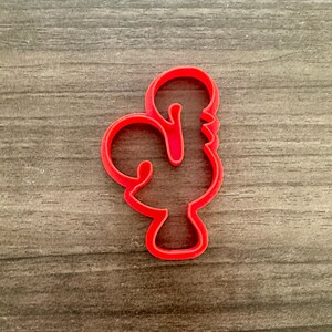 May include: A bright red cookie cutter in the shape of a rooster. The cutter is made of plastic and has a smooth, clean finish. The rooster design is detailed, with a curved body, a comb, and a beak. The cutter is designed for making cookies or other baked goods.