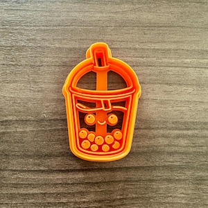 May include: An orange plastic cookie cutter in the shape of a bubble tea cup. The cutter features a straw, a smiling face, and several round boba pearls. The design is suitable for making cookies or other treats.