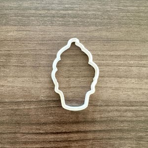 May include: A white plastic cupcake-shaped cookie cutter. The cutter has a detailed outline of a cupcake with a swirled frosting top and a cup-shaped base. The cutter is designed for baking and crafting.