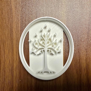 May include: White plastic cookie cutter featuring a stylized tree design within a circular frame. The tree has intricate branches and roots, with small diamond-shaped accents. The cutter is designed for baking and crafting.