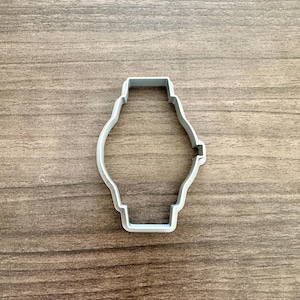 May include: A light gray cookie cutter in the shape of a watch. The cutter has a rectangular top and bottom with a rounded center. The cutter is made of plastic and sits on a wooden surface.