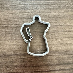 May include: A light gray plastic cookie cutter in the shape of a stovetop espresso maker, also known as a Moka pot. The cutter is on a wooden surface.