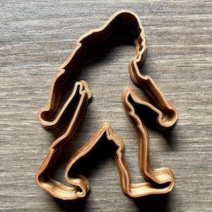 May include: A brown plastic cookie cutter shaped like a Bigfoot walking. The cookie cutter is detailed with a textured surface.