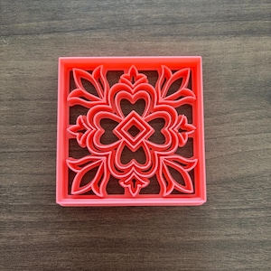May include: A square, coral-colored cookie cutter with a floral design. The cutter features a symmetrical pattern of leaves, hearts, and a central diamond shape. The design is raised, creating a detailed impression for baking.