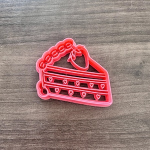 May include: A pink plastic cookie cutter in the shape of a slice of pie. The pie slice has a decorative crust, a strawberry, and rows of heart-shaped cutouts. The cutter is on a wooden surface.