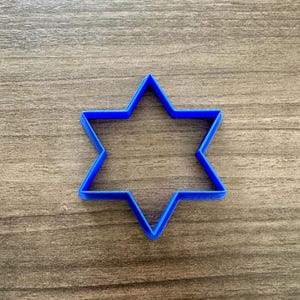 May include: A blue, six-pointed star-shaped cookie cutter. The cutter is made of plastic and sits on a wooden surface. The star has sharp points and a hollow center, perfect for baking themed cookies.