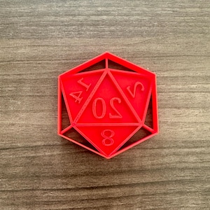 May include: A red, geometric D20 die-shaped cookie cutter. The numbers 4, 0, 8, and 5 are visible on the die. The cutter is made of plastic and is sitting on a wooden surface.