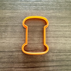 May include: An orange plastic cookie cutter in the shape of a spool of thread. The cutter has a rounded top and bottom with a rectangular center. The cutter is made of a bright orange plastic.