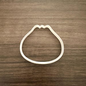 May include: A white plastic cookie cutter in the shape of a dumpling, with a wavy top edge. The cutter is set against a brown wooden surface.