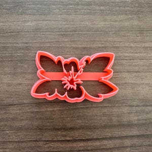 May include: A coral-colored floral cookie cutter. The cutter features a stylized flower design with curved petals and a central starburst shape. The cutter is made of plastic and is designed for baking.