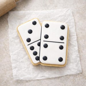 May include: Two domino-shaped cookies with white icing and black dot details. The cookies have a light brown, rectangular shape and are placed on a piece of white parchment paper. A rolling pin is visible in the background.