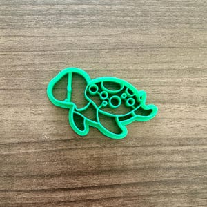 May include: A bright green turtle-shaped cookie cutter. The cutter is made of plastic and features a detailed design of a turtle with a shell and flippers. The cutter is designed for making cookies or other treats.