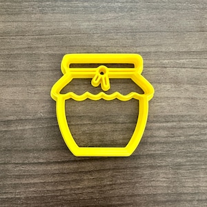 May include: A bright yellow plastic cookie cutter in the shape of a honey jar. The cutter has a wavy edge for the jar's opening and a small bow detail on the lid. The cutter is set against a gray wooden surface.