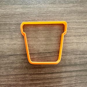 May include: An orange plastic cookie cutter in the shape of a flower pot. The cutter has a rectangular shape with a slightly flared top and a flat bottom. The cutter is made of a bright orange plastic.