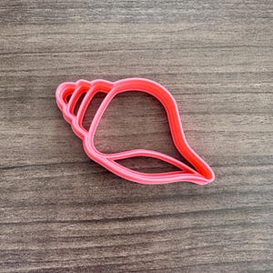 May include: A bright pink seashell-shaped cookie cutter. The cutter is made of plastic and has a detailed outline of a conch shell. The cutter is on a wooden surface.