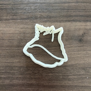 May include: A white plastic cookie cutter in the shape of a rose. The cutter has a detailed outline of the flower's petals and leaves. The cutter is on a brown wooden surface.