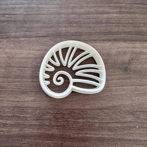 May include: A white plastic seashell-shaped cookie cutter. The cutter features a spiral design on one end and radiating lines on the other, resembling a nautilus shell. The cutter is designed for baking and crafting.