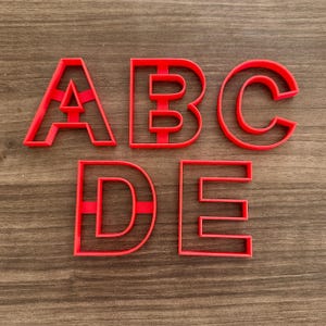 May include: Red plastic letter cookie cutters spelling out "ABCDE". The letters are in a sans-serif font and are arranged in a single row. The cutters are on a wooden surface.