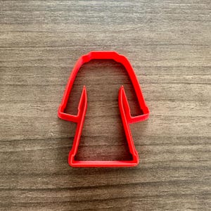 May include: A bright red cookie cutter in the shape of a robe or coat. The cutter is made of plastic and has a simple, clean design. The cutter is designed for making cookies or other treats.