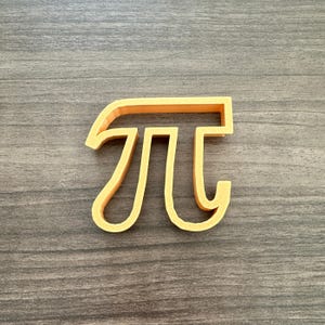 May include: A yellow-orange Pi symbol cookie cutter. The mathematical symbol is in a sans-serif font and is made of plastic. The cookie cutter is on a gray wooden surface.