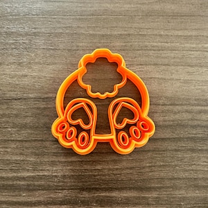 May include: An orange plastic cookie cutter in the shape of a bunny's rear end. The cutter features the bunny's fluffy tail and two feet with paw prints. The cutter is designed for making cookies or other treats.