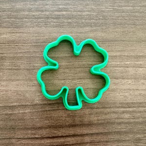 May include: A bright green four-leaf clover cookie cutter. The clover shape is made of plastic and sits on a wooden surface. The cookie cutter is a St. Patrick's Day themed item.