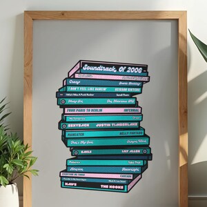 Könnte beinhalten: Gerahmter Druck mit einem Stapel von Büchern mit teal- und pinkfarbenen Buchrücken. Die Bücher tragen Songtitel und Künstlernamen aus dem Soundtrack von 2006. Der Rahmen ist aus hellem Holz.