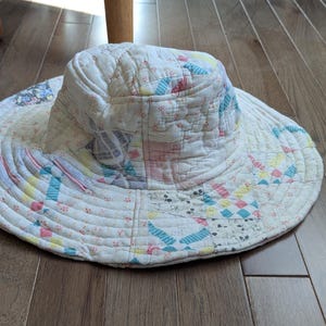 May include: A quilted sun hat with a wide brim, featuring a patchwork design in pastel colors. The hat is made of fabric and has a circular shape. The brim is approximately 14 inches in diameter.