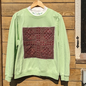 Op de afbeelding: Een lichtgroene sweatshirt met een vierkante, gewatteerde patch in bruintinten. De sweatshirt hangt aan een houten hanger. Een wit ondershirt met bloemenborduurwerk is zichtbaar bij de halslijn. De achtergrond is een houten muur.