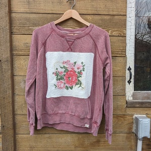 May include: A maroon sweatshirt with a white square patch featuring a floral design in shades of pink, green, and white.