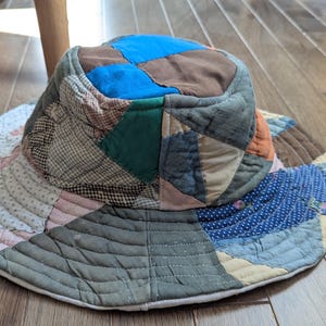 Può includere: Un cappello da sole patchwork con tesa larga. Il cappello presenta una varietà di motivi e colori di tessuto, tra cui blu, verde, marrone e rosa. Il cappello è realizzato in tessuto trapuntato e ha una corona arrotondata.