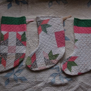 May include: Three quilted Christmas stockings with a patchwork design. Each stocking features a green cuff, a pink band, and a white body with red and green star and geometric patterns. They hang on wooden dowels.
