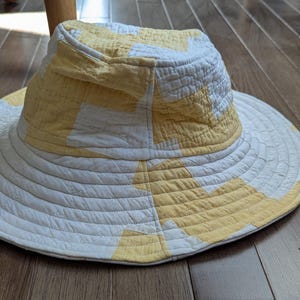May include: A wide-brimmed sun hat with a quilted design. The hat features a white and yellow color scheme, with the crown and brim displaying a patchwork pattern. The brim is wide and offers ample shade. The hat is made of a soft, likely cotton, material.