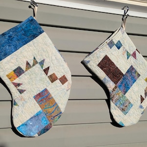 May include: Two handmade Christmas stockings with quilted patchwork designs. One stocking features blue and white fabric with geometric shapes, while the other has a mix of brown, blue, and white patterns. Both have blue cuffs and are hanging on a gray wall.