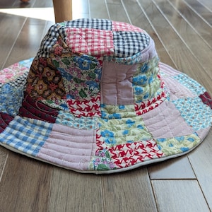 Op de afbeelding: Een patchwork zonnehoed met een brede rand. De hoed heeft een verscheidenheid aan stofpatronen, waaronder ruitjes, bloemen en effen kleuren in rood, blauw, roze en zwart. De hoed is gemaakt van gewatteerde stof en ligt op een houten vloer.