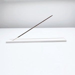 Könnte beinhalten: Ein rechteckiger weißer Räucherstäbchenhalter mit einem einzelnen braunen Räucherstäbchen darauf.