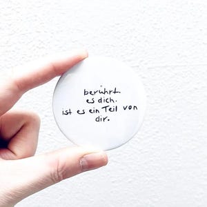 Könnte beinhalten: Ein weißer, runder Knopf mit handgeschriebenem schwarzem Text. Der Text, auf Deutsch, lautet: "berührt. es dich. ist es ein Teil von dir." Der Knopf wird zwischen zwei Fingern vor einem weißen Hintergrund gehalten.