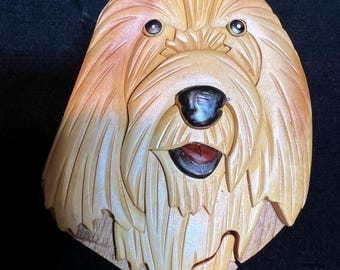 May include: A wooden puzzle box shaped like a dog's head. The dog has brown fur, black eyes, and a black nose. The box is made of light wood and has a detailed carving of the dog's fur.